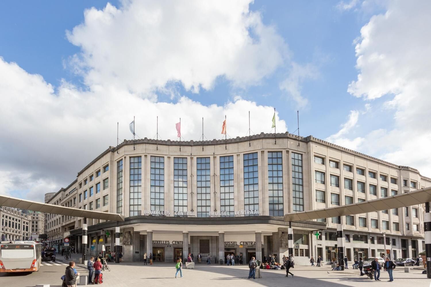 A large, historic building with multiple flags, a central entrance, and a spacious plaza with people and vehicles.