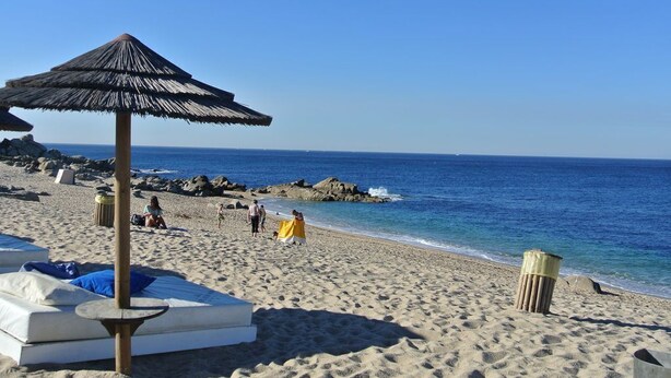 A beach with a thatched umbrella, a white lounge chair, and a mattress on the sand.