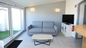 A modern living room with a grey sofa, a TV, a small table, and a window with a view of a grassy area.
