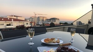 A table with a plate of food, two glasses of wine, and a bottle of wine on a rooftop terrace with a city view.
