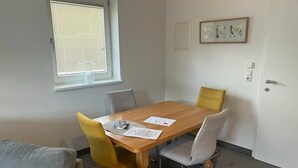 Una habitación pequeña y bien iluminada con una mesa de madera, cuatro sillas, un ventanal con persianas, una puerta y un cuadro en la pared.