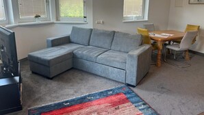 Una sala de estar con un sofá seccional gris, una mesa de comedor de madera con sillas, y una alfombra roja y azul.