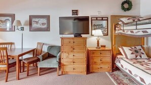 Ein gemütliches Zimmer mit Holzboden, einem Fernseher, einem Schreibtisch mit Stühlen, einem Sofa, einem Bett mit Patchworkdecke und einer Kommode.