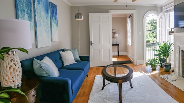 A living room with a blue sofa, a glass coffee table, and a fireplace. There are potted plants and a TV mounted on the wall. - Luxury on Main / North (Richmond)