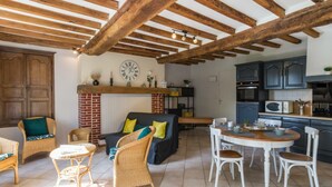 A spacious living area with a fireplace, wooden beams, wicker furniture, and a kitchen with dark cabinets.