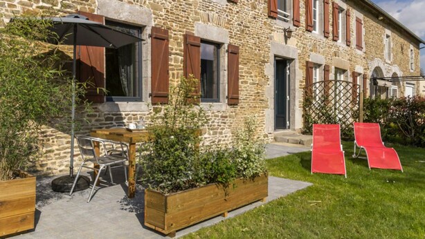 A stone building with wooden shutters, a patio with a table and chairs, and a garden area with red chairs.