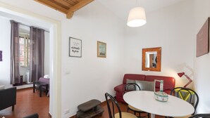 Uma área de refeições com uma mesa redonda, cadeiras e um banco. Há um sofá vermelho e uma mesinha com um abajur.