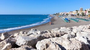Ein Strand mit felsigem Vordergrund, klarem blauem Meer und Gebäuden im Hintergrund.