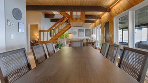 Una sala da pranzo spaziosa con un lungo tavolo in legno e sedie in vimini, una scala in legno e ampie finestre che offrono viste sull'esterno.
