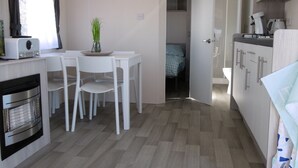 Uma cozinha compacta com micro-ondas, fogão e pia. Há uma área de refeições com mesa e cadeiras, e uma pequena planta sobre a mesa. Um quadro está pendurado na parede acima da área de refeições.