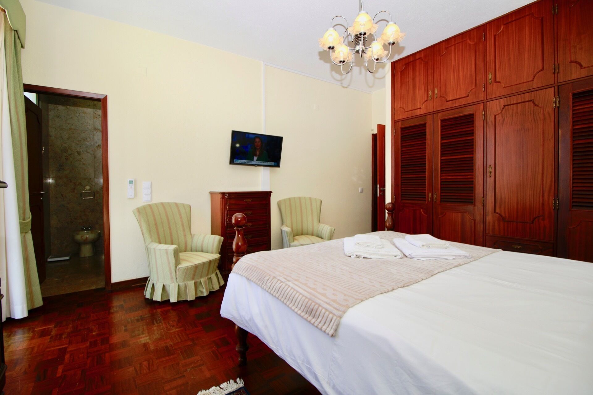A hotel room with a large bed, two armchairs, a wooden wardrobe, a television on the wall, and a bathroom with a marble wall and a sink.