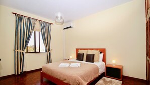 A bedroom with a wooden bed, a nightstand, a lamp, and a view of the outdoors through a window with curtains. - Villas Quinta da Lapa 2. Villa. Heated pool. WiFi (Câmara de Lobos)