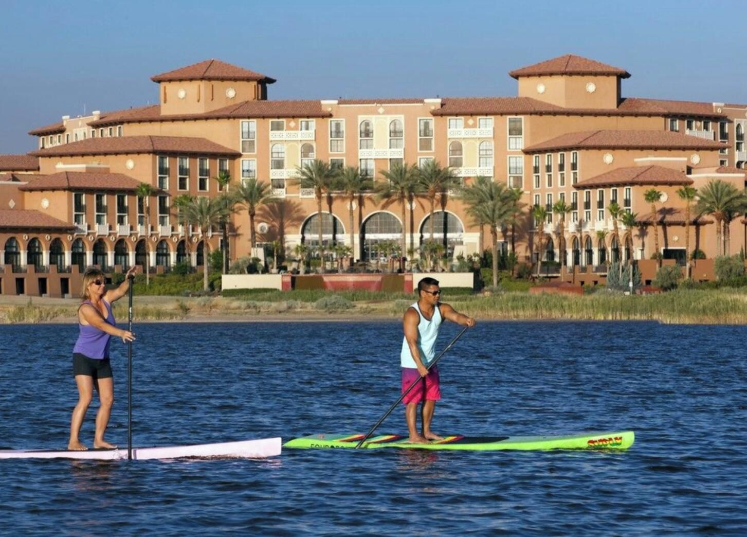 Two people paddleboarding on a calm body of water with a large hotel building in the background.