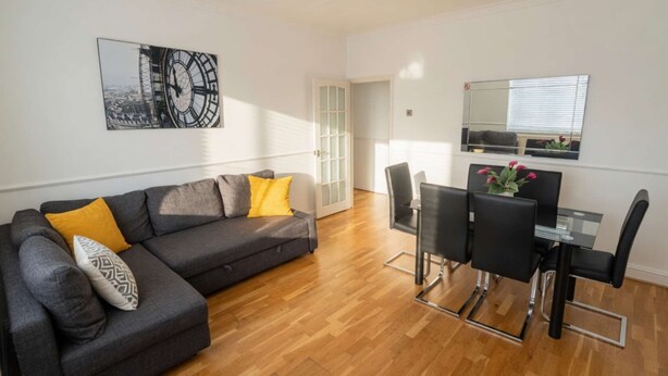 Een moderne woonkamer met een grijze bank, gele en gemêleerde kussens, een eethoek met een glazen tafel en zwarte stoelen, en een grote wandklok.