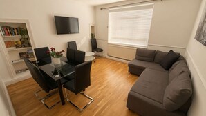 Een moderne woonkamer met een eethoek, een grijze bank, een flatscreen televisie en een boekenkast.