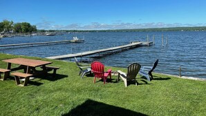 Area picnic sul lago con panche in legno e poltrone Adirondack, un molo che si protende nell'acqua e un cielo azzurro limpido.