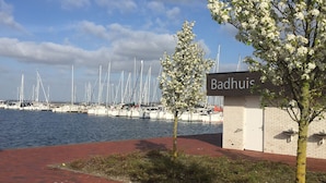 Un port de plaisance avec des bateaux amarrés, une enseigne indiquant « Badhuis », et un chemin pavé de briques menant à l’eau.