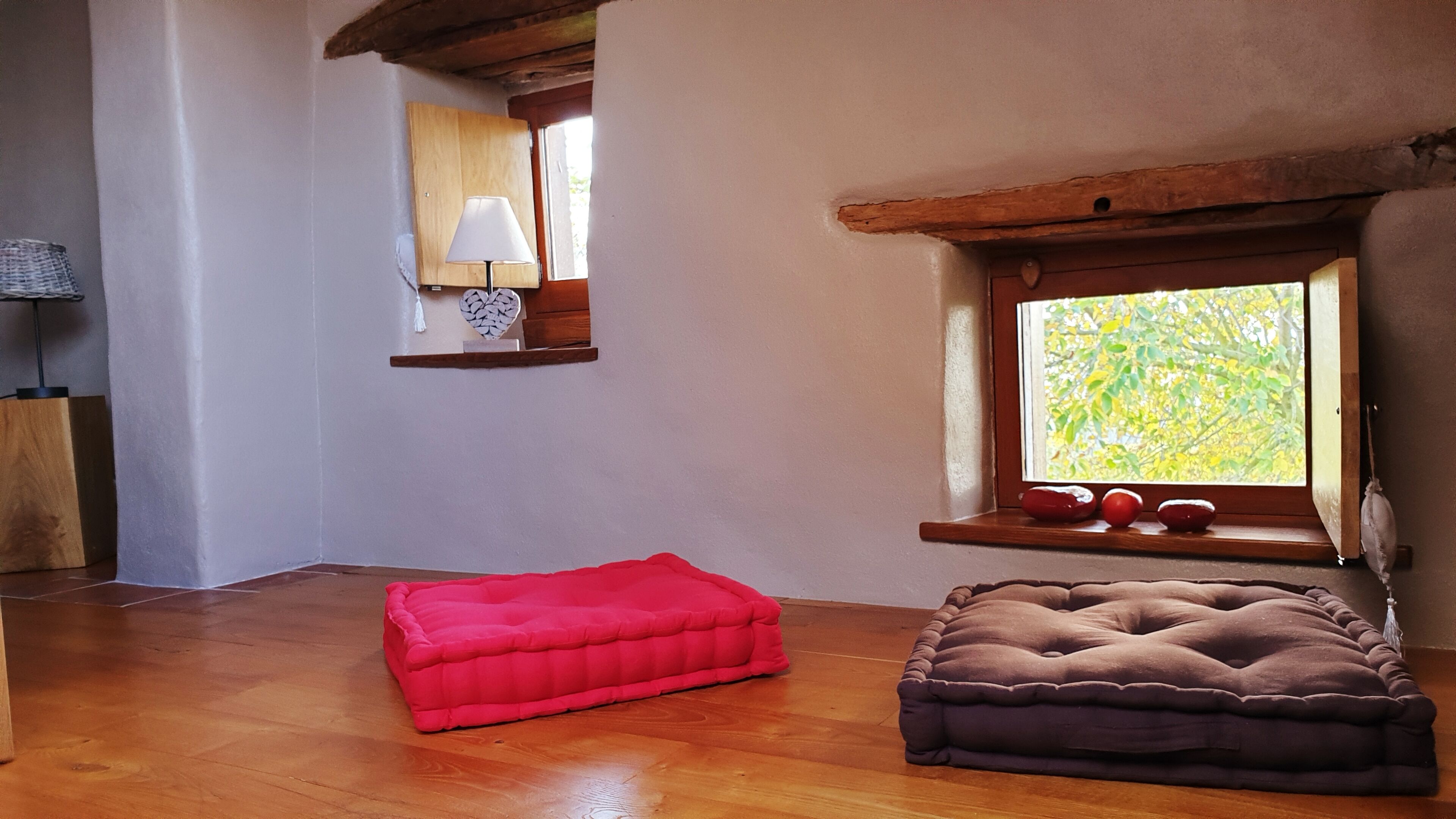 Una habitación con piso de madera, un cojín rojo, un cojín marrón, una ventana con vista a follaje y una repisa con una lámpara.