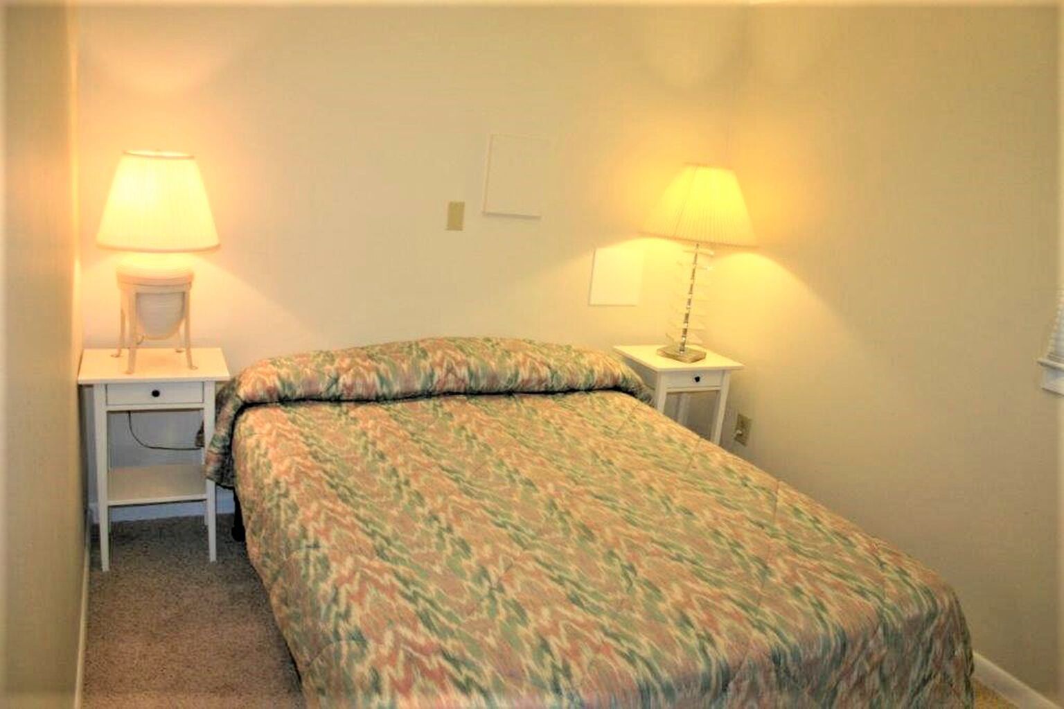 A small, single-bed room with a patterned bedspread, two bedside tables with lamps, and a chair.