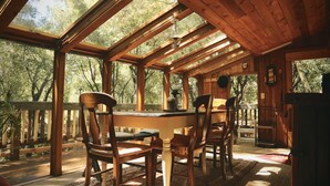 A wooden deck with a dining table and chairs, surrounded by large windows and a view of trees. - NEW: Elegant creekside cabin with waterfall (Idyllwild-Pine Cove)