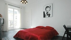 Une chambre à coucher avec un grand lit, une chaise, un bureau et une fenêtre donnant sur des immeubles.