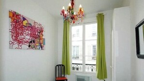 Une pièce comprenant un lit, une chaise, un tableau abstrait aux couleurs vives, une fenêtre avec des rideaux et un lustre.