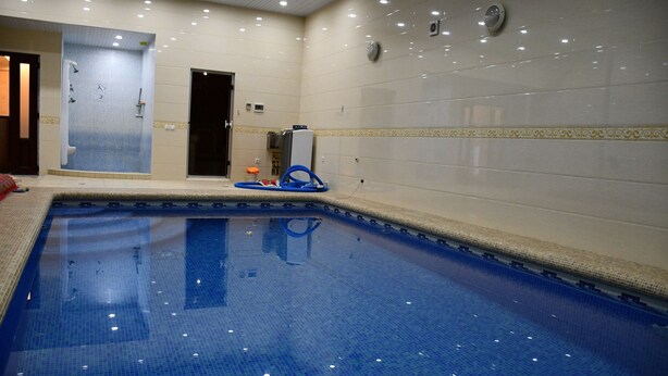 Una piscina interior con suelo y paredes de baldosas, zona de duchas y una puerta que da a otra habitación.