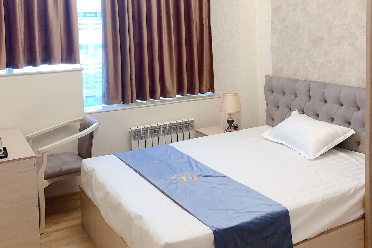 Une chambre d’hôtel avec un lit, un bureau, une chaise, un radiateur et une fenêtre avec des rideaux.