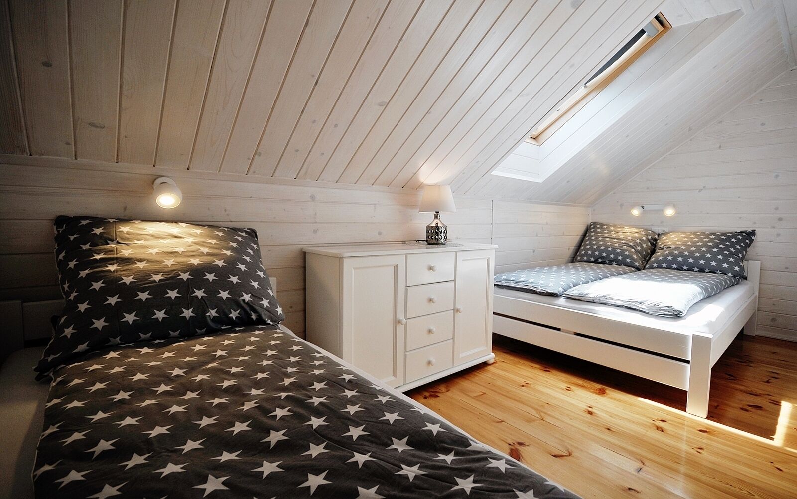 Une chambre avec un plafond en pente, un puits de lumière, un lit avec une literie à motifs d’étoiles, une table de chevet avec une lampe et une commode blanche.