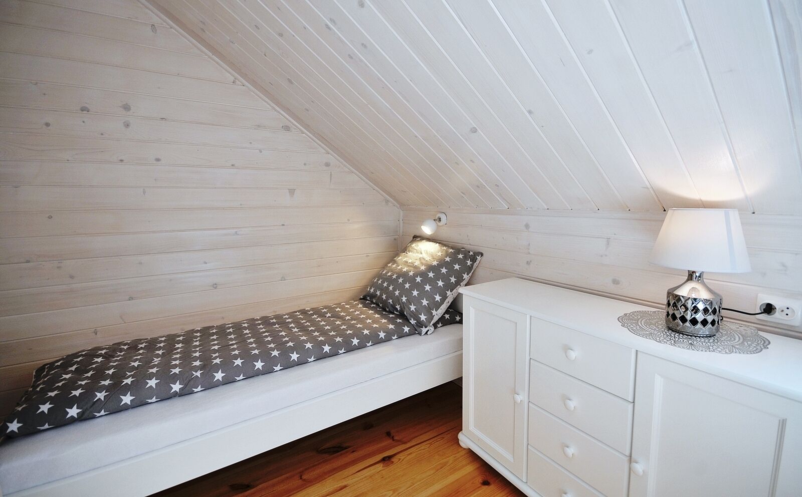 A small, slanted-roofed room with a single bed, a bedside table with a lamp, and a white dresser.