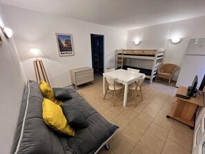 Un espace de vie compact comprenant un canapé, une table, un lit superposé, un petit meuble et un réfrigérateur.