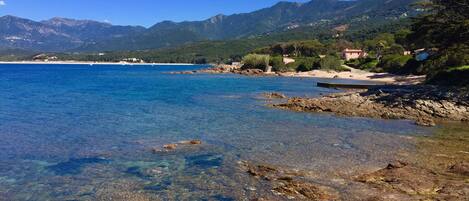 A coastal landscape with clear waters, rocky shore, and distant mountains.