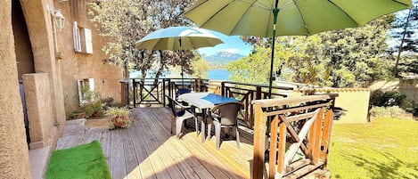 An outdoor patio with wooden tables and chairs, a green umbrella, and a view of the sea.
