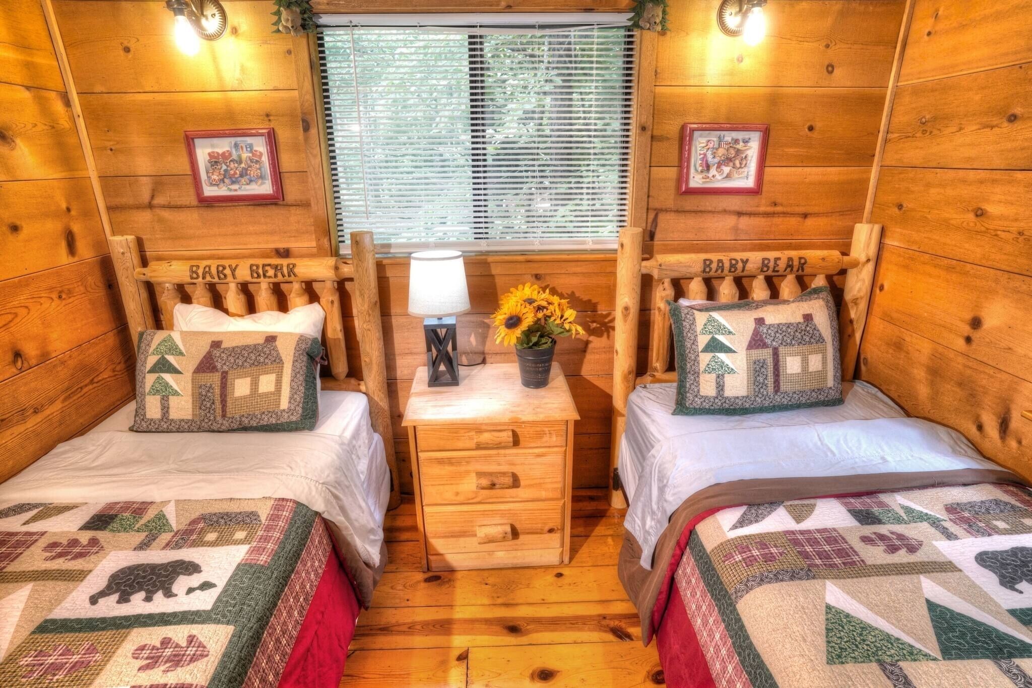 A cabin-style room with two beds labeled 'BABY BEAR,' a small table with a lamp, and a window with blinds.