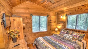 A wooden cabin bedroom with a bed, two windows, a bedside lamp, a TV, and a small table with a plant. - Three Bears Cabin- Enjoy the scenery around you with a peaceful view (Sautee Nacoochee)
