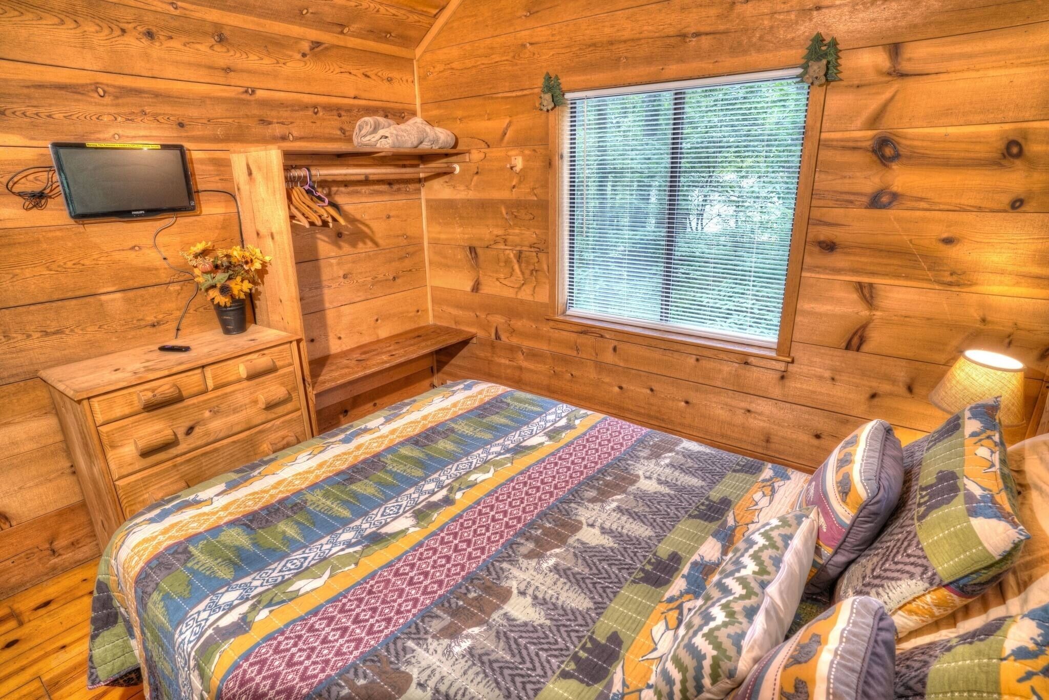 A wooden cabin room with a bed, a TV mounted on the wall, a window with blinds, and a small shelf with a plant.