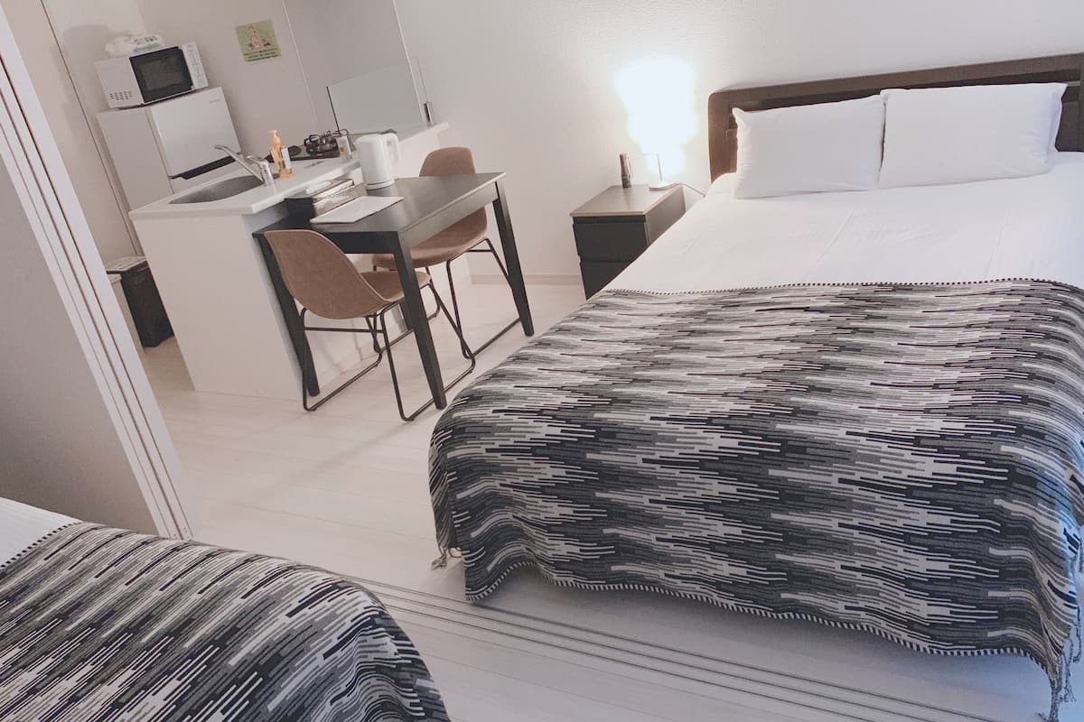 A hotel room with a bed, a desk, and a chair. There is a microwave and a kettle on the desk.