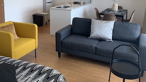 Una sala moderna con un sofá gris, un sillón amarillo y un sofá estampado. Se ve un comedor con una mesa y sillas al fondo.