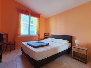 Een slaapkamer met een bed, nachtkastje, stoel en raam met gordijnen.