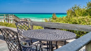 Een houten terras met een ronde metalen tafel en stoelen, uitkijkend op een turquoise oceaan.