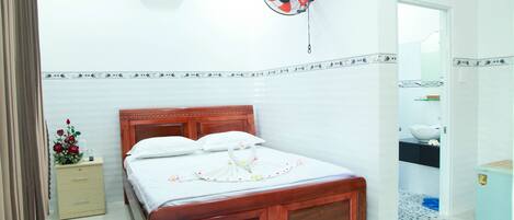 Una cama bien hecha con cabecera de madera, una mesita de noche con un jarrón de flores, un ventilador de pared y un refrigerador pequeño.