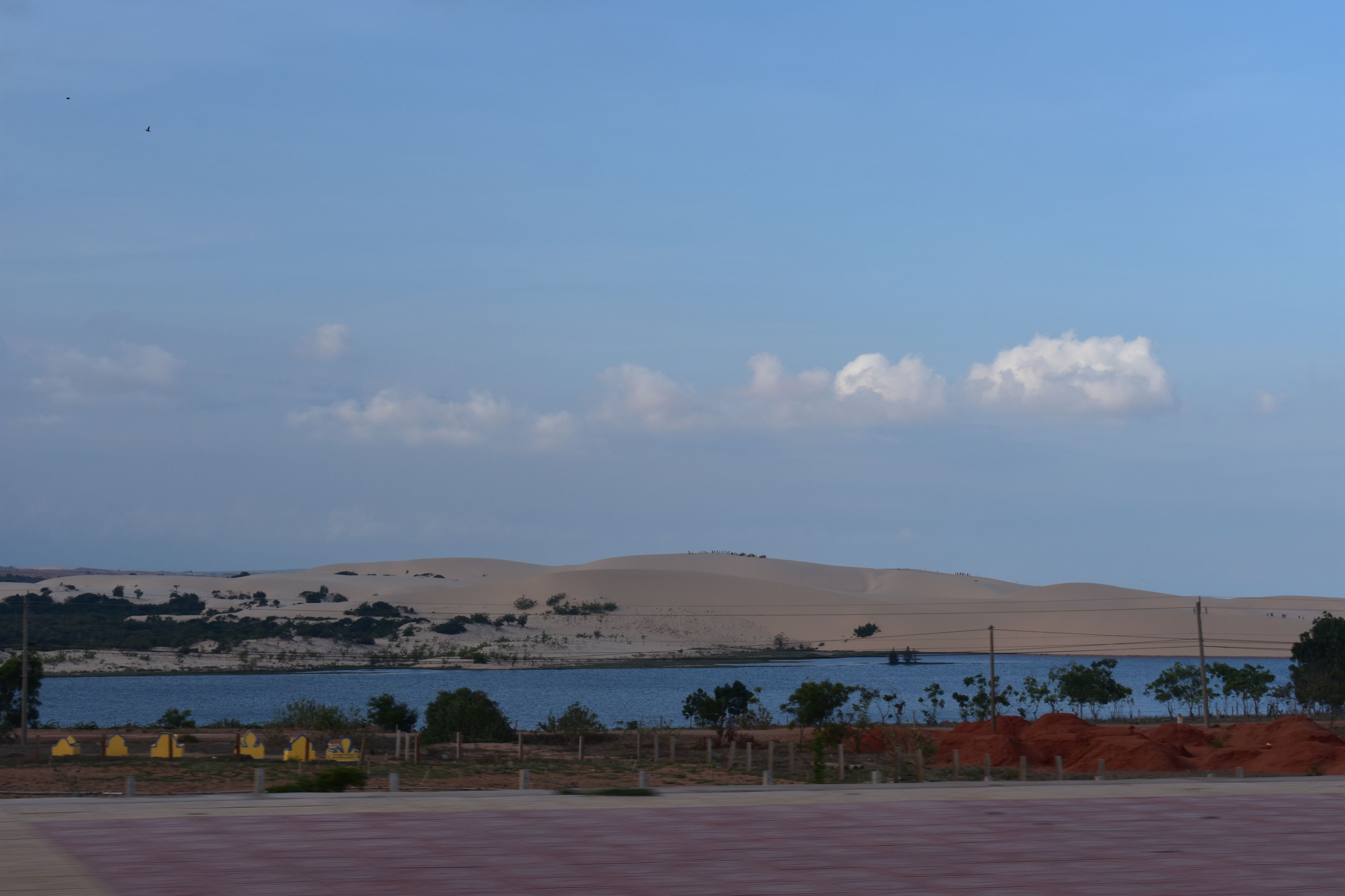 A desert landscape with sand dunes, a body of water, and sparse vegetation.
