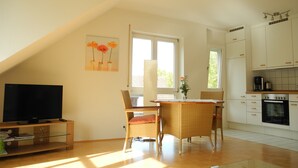 Eine moderne Küche mit weißen Schränken, ein Essbereich mit Korbstühlen, einem Flachbildfernseher und einem Blumenbild an der Wand.