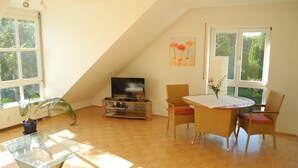 Ein Zimmer mit Fernseher auf einem Holzständer, einem Esstisch mit Stühlen, einer Topfpflanze und großen Fenstern.