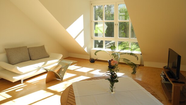 Ein modernes Wohnzimmer mit schrägen Wänden, einem weißen Sofa, einem Glastisch und einem Fernseher auf einem Holzboden.