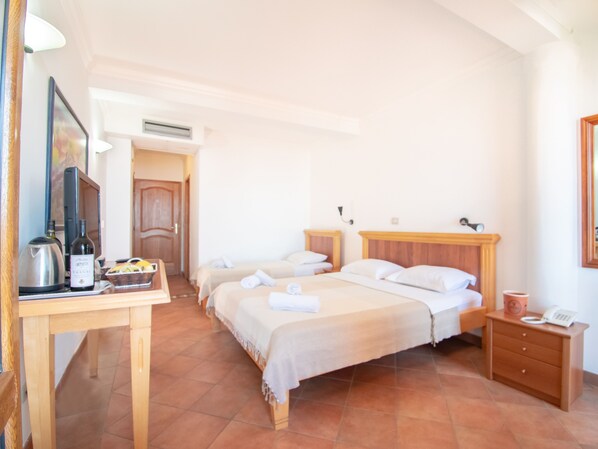 A hotel room with two beds, a wooden nightstand, a desk with a kettle and wine bottle, and a telephone.
