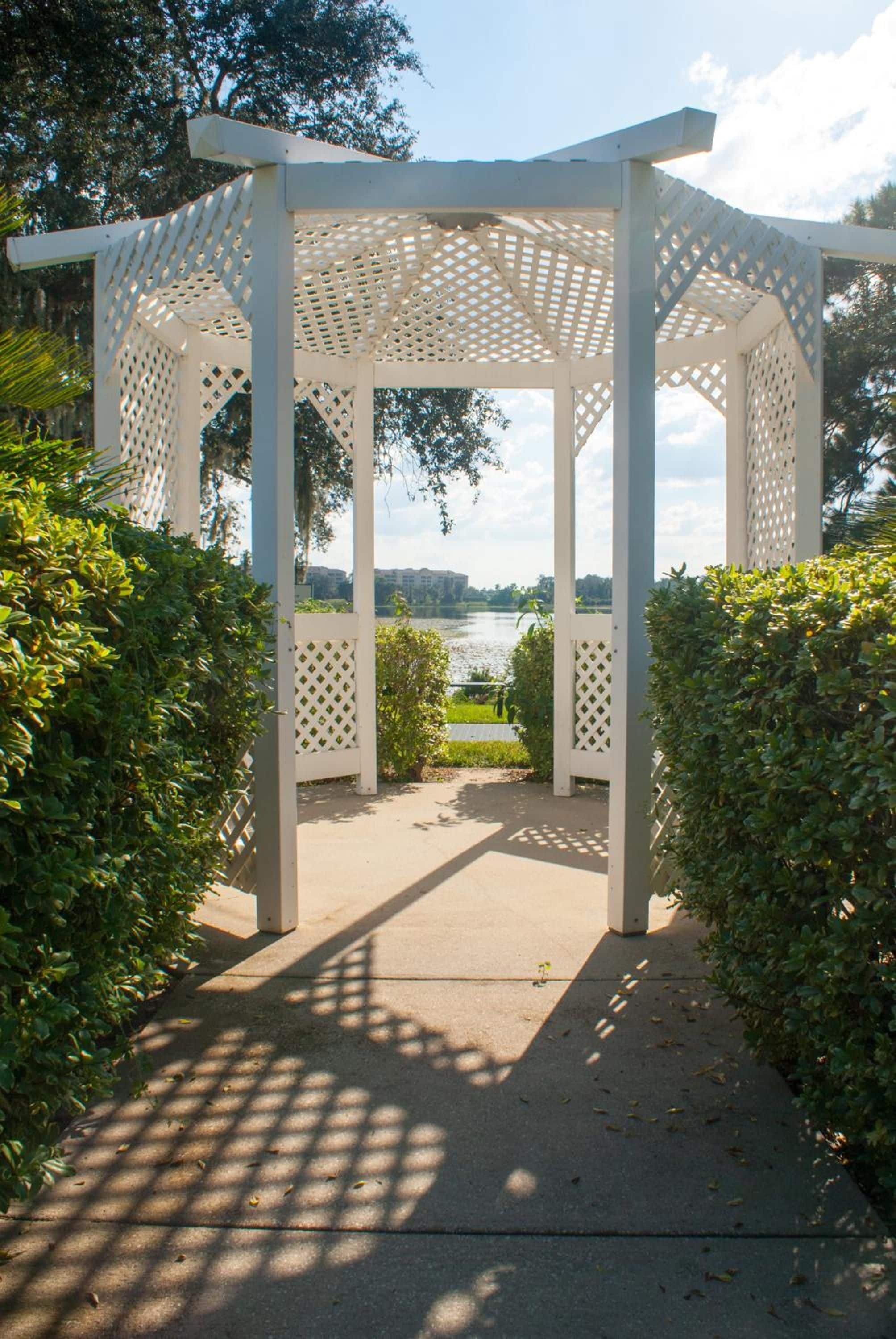 A white pergola structure with a lattice design, surrounded by greenery and leading to a view of a body of water.