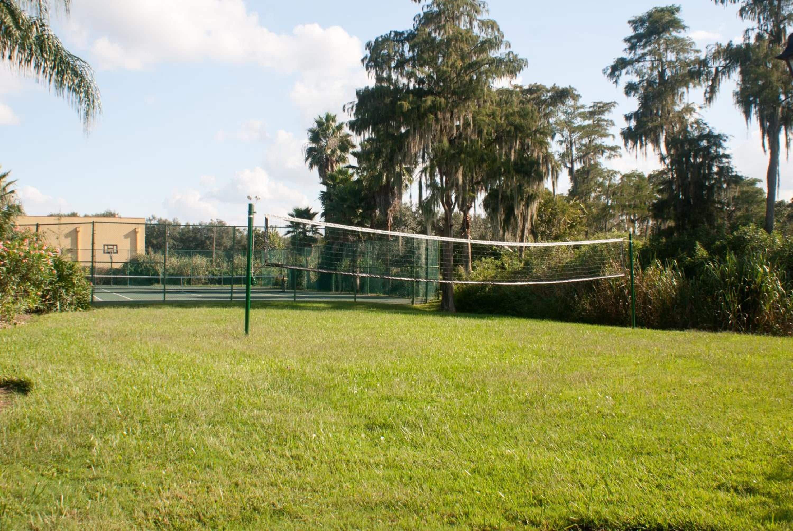 Uma área gramada com uma rede de voleibol, cercada por árvores e um edifício ao fundo.