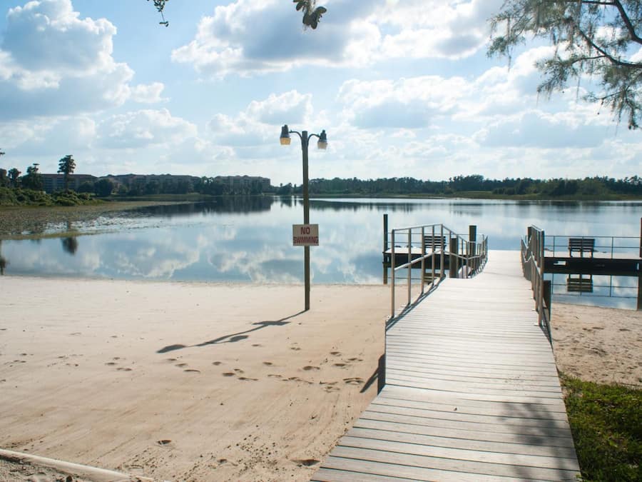 Une jetée en bois menant à un lac paisible, avec un panneau indiquant « BAIGNADE INTERDITE », entourée d’arbres et sous un ciel dégagé.
