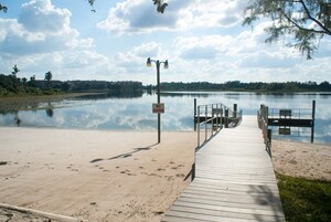 Un muelle de madera que lleva a un lago tranquilo con un letrero de 'NO NADAR', rodeado de árboles y un cielo despejado.